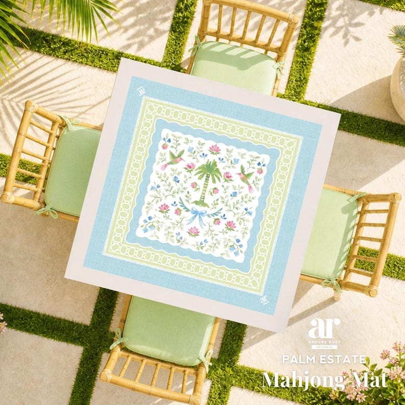 May include: A square Mahjong mat with a light blue border and a central design featuring a palm tree, hummingbirds, and floral elements. The mat is set on a table with bamboo chairs and green cushions.