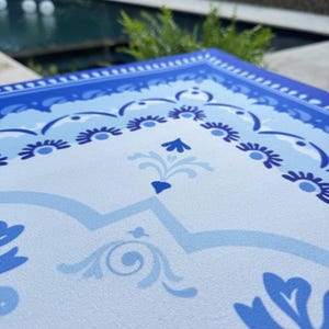 May include: A close-up of a blue and white decorative placemat with a floral design. The placemat has a border of dark blue and light blue floral patterns. The center of the placemat has a light blue background with a repeating floral design.