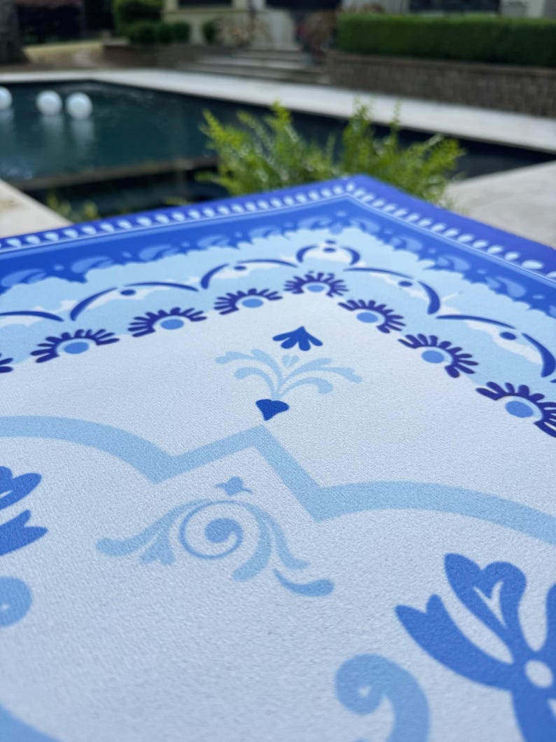 May include: A close-up of a blue and white decorative placemat with a floral design. The placemat has a border of dark blue and light blue floral patterns. The center of the placemat has a light blue background with a repeating floral design.