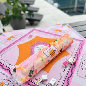 Pu&ograve; includere: Un set di giochi a tema Charleston rosa e arancione con un pezzo di tessuto arrotolato con illustrazioni di bottiglie di champagne e la scritta "Let's Rock & Roll". Il set include tessere di gioco e una superficie rosa a motivi.