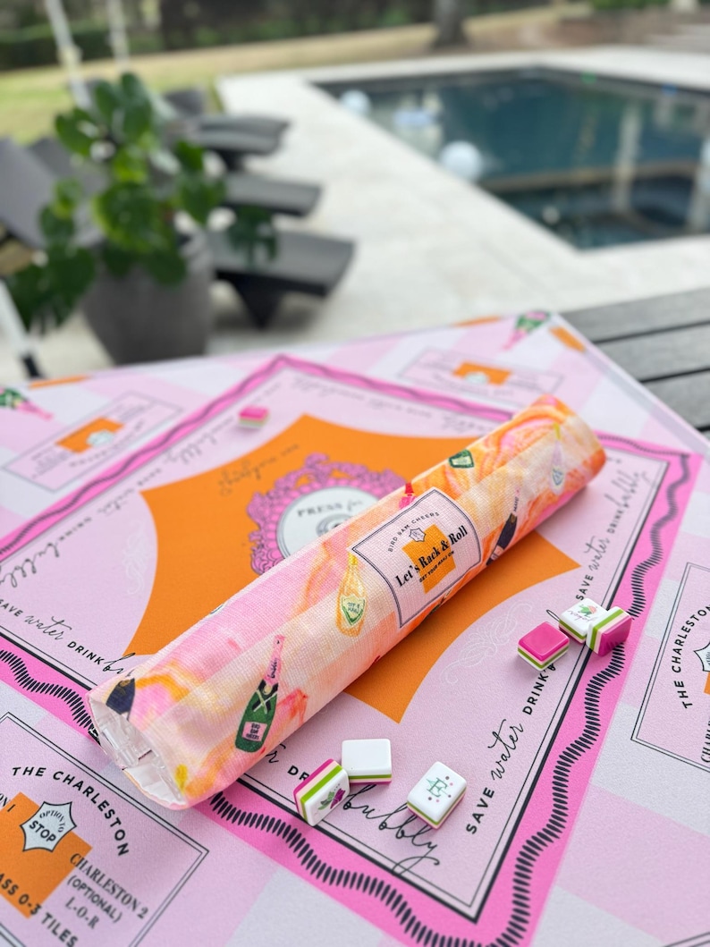 Pu&ograve; includere: Un set di giochi a tema Charleston rosa e arancione con un pezzo di tessuto arrotolato con illustrazioni di bottiglie di champagne e la scritta "Let's Rock & Roll". Il set include tessere di gioco e una superficie rosa a motivi.