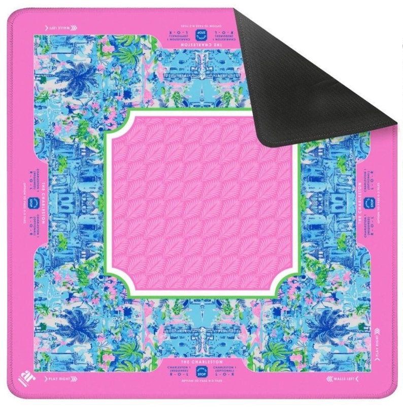 Pode incluir: Um mousepad retangular rosa com um design floral e arquitet&ocirc;nico colorido. O centro apresenta um padr&atilde;o de folhas rosa dentro de uma moldura com bordas brancas. As bordas s&atilde;o adornadas com ilustra&ccedil;&otilde;es florais e de edif&iacute;cios azuis e verdes, com o texto "THE CHARLESTON".