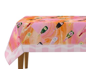 May include: A pink and orange tablecloth featuring a pattern of champagne bottles. The tablecloth has a white and pink striped border and is draped over a wooden table. The bottles are in various colors, including green, gold, and black.