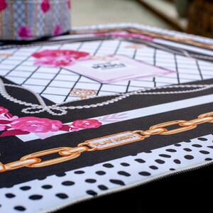 May include: A decorative mat with a black and white design, featuring gold chain links, pink roses, and a perfume bottle illustration. The mat has the words "PLAY RIGHT" on a gold plate.