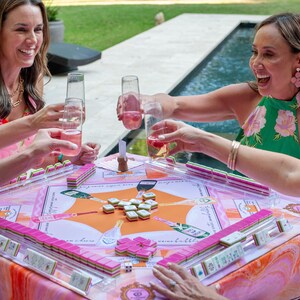 May include: A group of people are gathered around a table playing mahjong. The table is covered with a pink and orange patterned cloth, and the mahjong tiles are pink. The people are holding glasses of pink drinks.