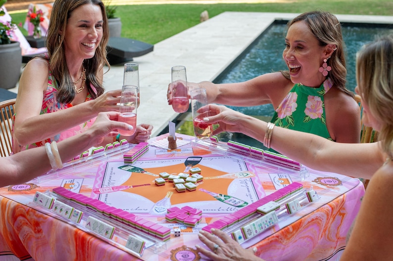 May include: A group of people are gathered around a table playing mahjong. The table is covered with a pink and orange patterned cloth, and the mahjong tiles are pink. The people are holding glasses of pink drinks.