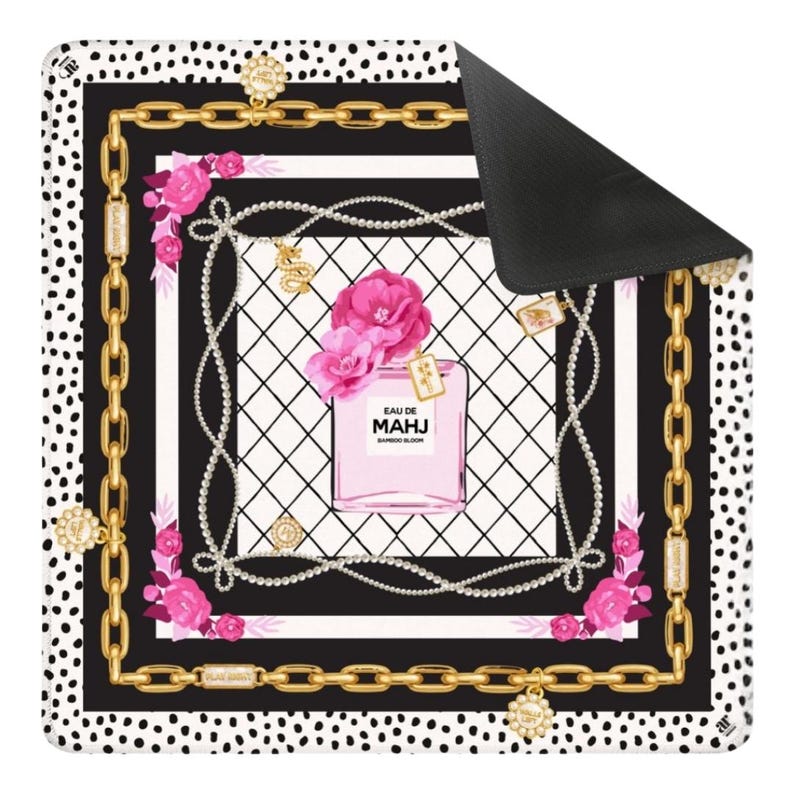 Op de afbeelding: Een decoratieve mat met een parfumflesontwerp. De mat heeft een zwart-witte stippelrand, een gouden kettingontwerp en een centraal beeld van een roze parfumfles met de tekst "EAU DE MAHJ BAMBOO BLOOM".