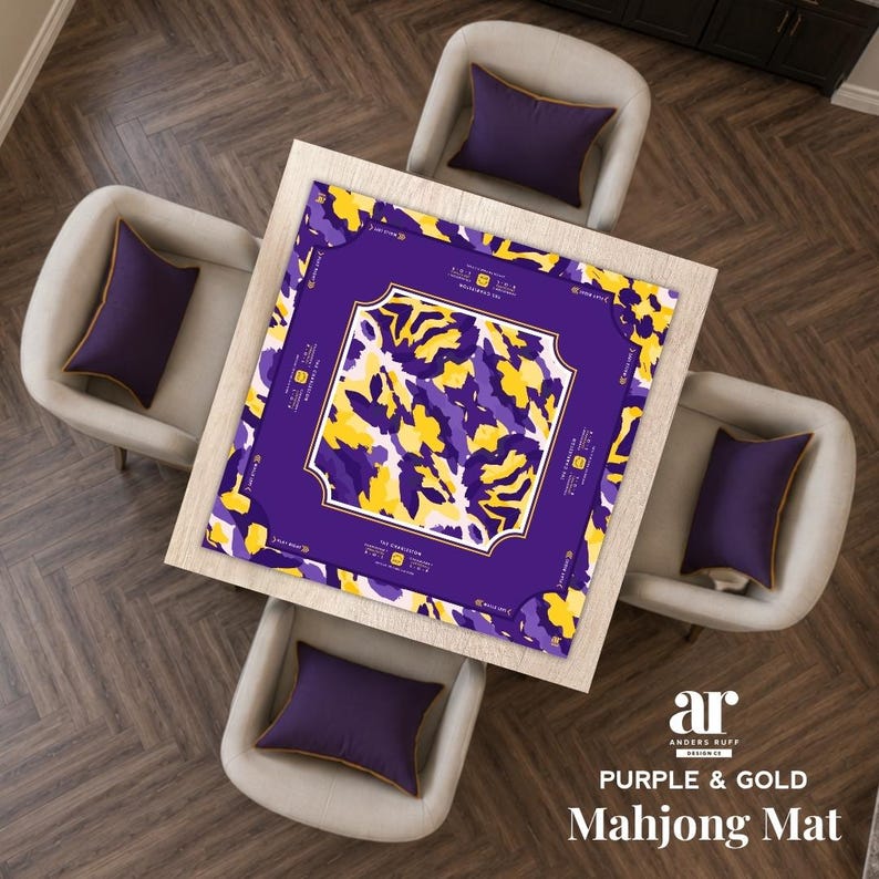 May include: A square mahjong mat featuring a central abstract pattern of purple, yellow, and white shapes. The mat has a solid purple border with white text and decorative line accents. The text reads The Charleston and Anders Ruff Design Co.