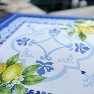 K&ouml;nnte beinhalten: Ein rechteckiger Tischset mit blauem und wei&szlig;em Blumendesign. Das Tischset hat einen Rand mit blauen Blumenmustern und ein zentrales Design mit Zitronen, Bl&auml;ttern und wei&szlig;en Bl&uuml;ten. Die Farbpalette umfasst Blau-, Gelb- und Gr&uuml;nt&ouml;ne.