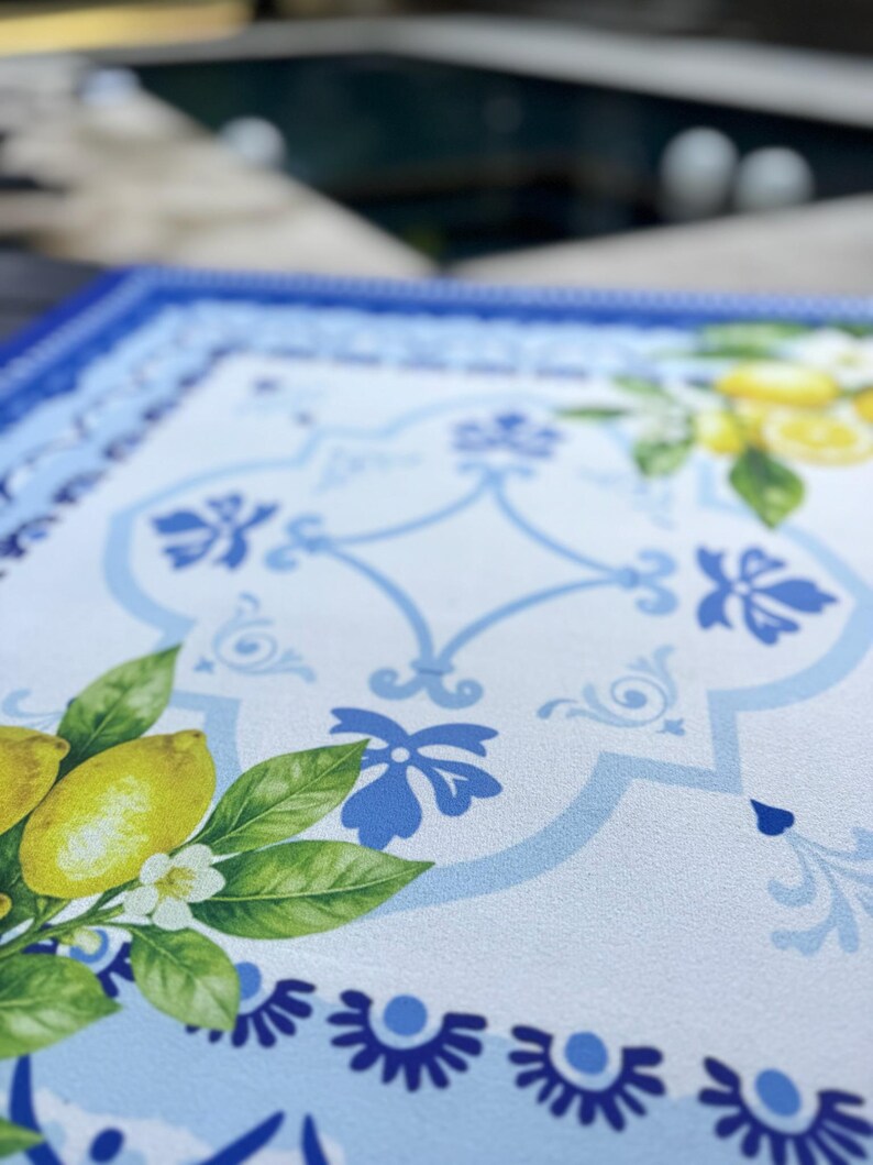 K&ouml;nnte beinhalten: Ein rechteckiger Tischset mit blauem und wei&szlig;em Blumendesign. Das Tischset hat einen Rand mit blauen Blumenmustern und ein zentrales Design mit Zitronen, Bl&auml;ttern und wei&szlig;en Bl&uuml;ten. Die Farbpalette umfasst Blau-, Gelb- und Gr&uuml;nt&ouml;ne.