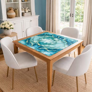 May include: A square table mat featuring a vibrant teal, blue, and white marbled pattern with gold-toned veining accents. The mat is positioned flat on the center of a wooden dining table, displaying a smooth surface with a detailed abstract design.