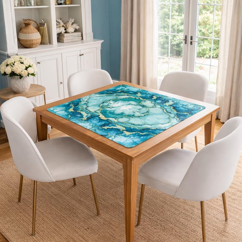 May include: A square table mat featuring a vibrant teal, blue, and white marbled pattern with gold-toned veining accents. The mat is positioned flat on the center of a wooden dining table, displaying a smooth surface with a detailed abstract design.
