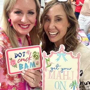 May include: Two women holding photo props with text. One prop says "DOT crack BAM" with floral and dragon designs. The other says "get your MAHJ ON" with a hummingbird and palm tree. Both props have pink and blue accents.