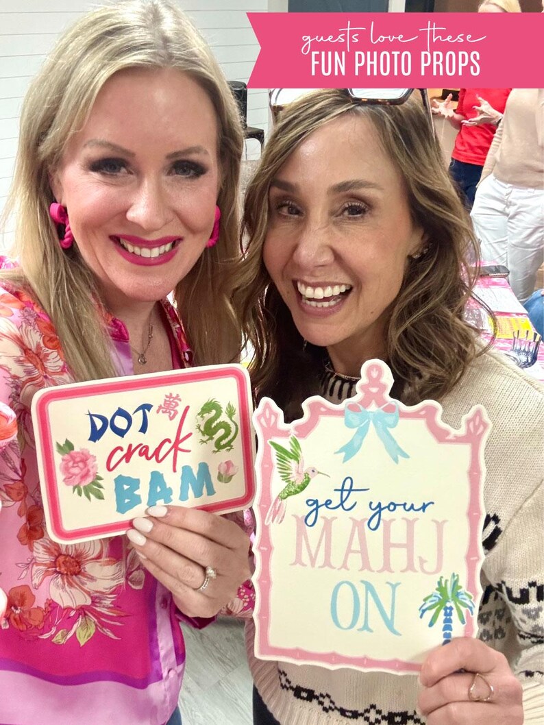 May include: Two women holding photo props with text. One prop says "DOT crack BAM" with floral and dragon designs. The other says "get your MAHJ ON" with a hummingbird and palm tree. Both props have pink and blue accents.