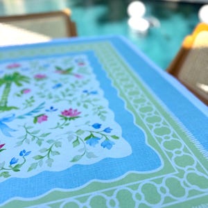 May include: A rectangular tablecloth with a light blue border and a white center featuring a floral and palm tree design. The tablecloth has a scalloped edge and a green decorative border.