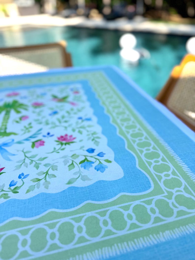 May include: A rectangular tablecloth with a light blue border and a white center featuring a floral and palm tree design. The tablecloth has a scalloped edge and a green decorative border.