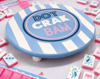 May include: A light blue and white striped wooden game board with the text "DOT CRAK BAM" in a circle. The board is surrounded by pink and white game tiles, and pink game pieces.