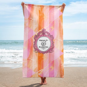 May include: A beach towel with a vibrant design of pink, orange, and white stripes and geometric shapes. The towel features the text "PRESS for MAHJONG" within a decorative pink frame. The towel is held up on a sandy beach.