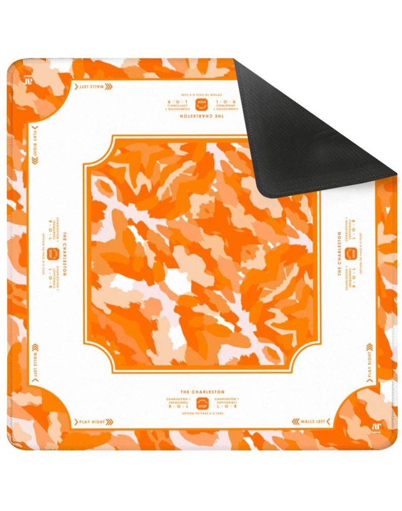 May include: A square, orange and white mat with a black underside. The mat features an abstract orange pattern and text that reads "THE CHARLESTON" and "WALLS LEFT" and "PLAY RIGHT". The mat has rounded corners.