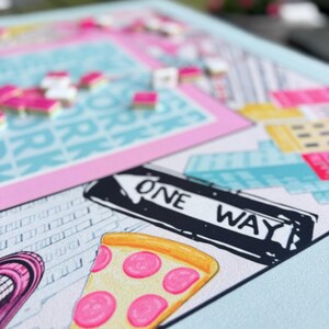 May include: A colorful board game with a New York City theme. The game board features a slice of pizza, a one-way street sign, and city buildings. Pink, blue, yellow, and white are the dominant colors. Game pieces are scattered across the board.