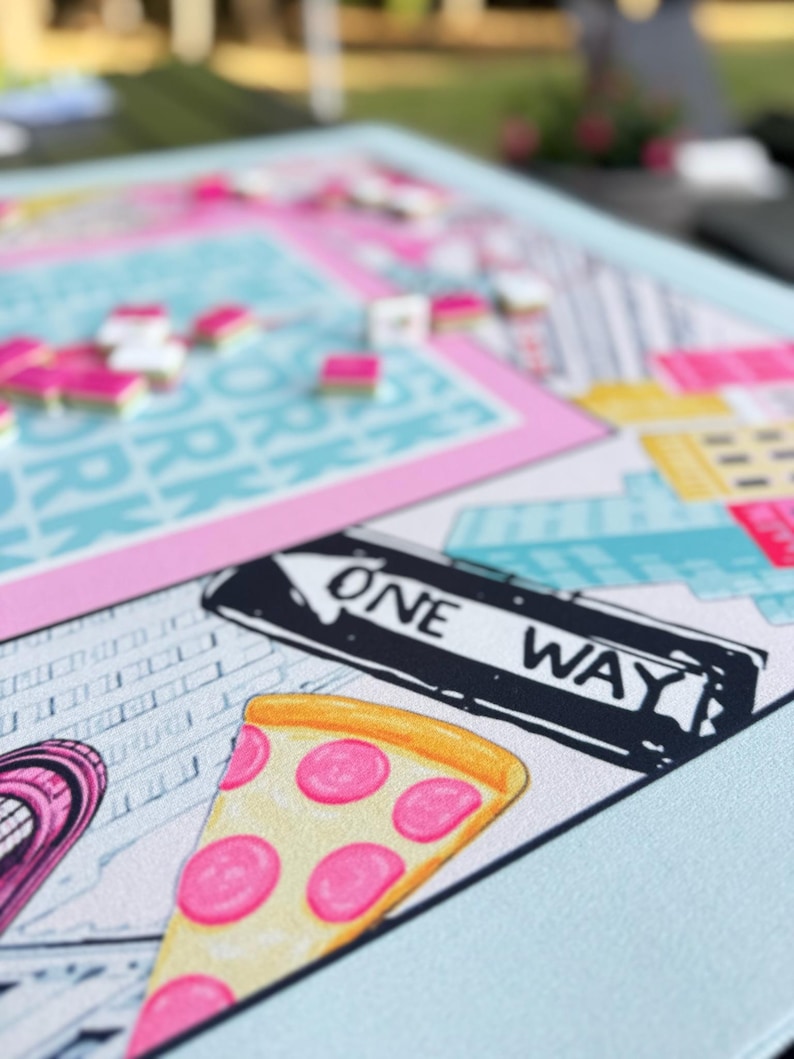 May include: A colorful board game with a New York City theme. The game board features a slice of pizza, a one-way street sign, and city buildings. Pink, blue, yellow, and white are the dominant colors. Game pieces are scattered across the board.