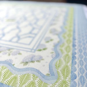 May include: Close-up of a patterned paper with a watercolor design. The design features a light blue and white background with a geometric pattern, green fern-like leaves, and blue wavy lines. The overall theme is floral and botanical.