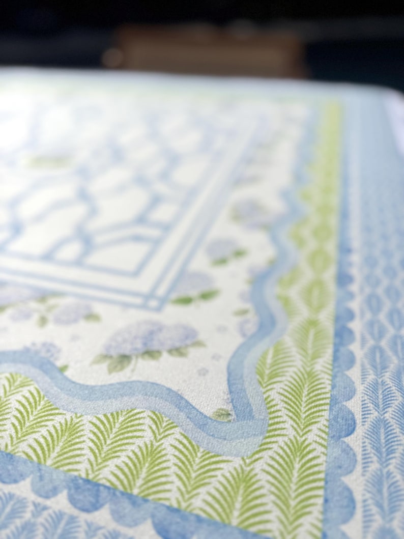 May include: Close-up of a patterned paper with a watercolor design. The design features a light blue and white background with a geometric pattern, green fern-like leaves, and blue wavy lines. The overall theme is floral and botanical.