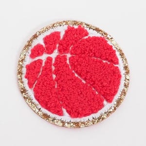 May include: A round, embroidered patch featuring a red fruit slice design with a white border. The fruit's texture is fluffy, and the outer edge is embellished with gold glitter.