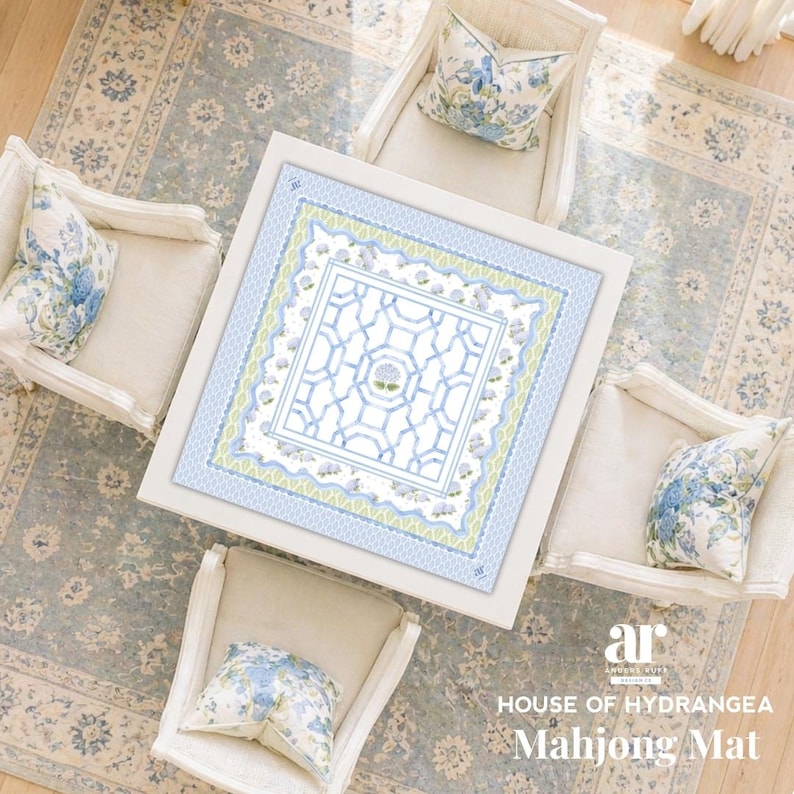 May include: Overhead view of a white square table with a blue and white patterned Mahjong mat. Four cream-colored armchairs surround the table, each with a floral patterned pillow. The rug beneath is blue with a floral design. Text reads "HOUSE OF HYDRANGEA Mahjong Mat."