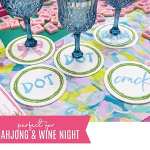 May include: White coasters with green bamboo-style borders and blue text reading "DOT", "BAM", and "crack" are under blue wine glasses. The coasters are on a colorful floral patterned surface. Pink Mahjong tiles are in the background.