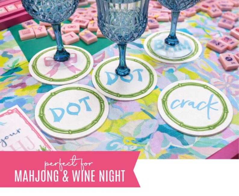 May include: White coasters with green bamboo-style borders and blue text reading "DOT", "BAM", and "crack" are under blue wine glasses. The coasters are on a colorful floral patterned surface. Pink Mahjong tiles are in the background.