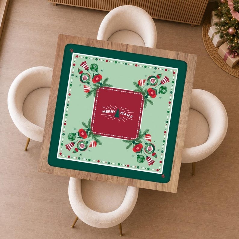 May include: A square table mat with a dark green border and a light green centre featuring red and green Christmas ornaments, pine branches, and a central red square with the white text MERRY MAHJ and a small tree icon in the middle.