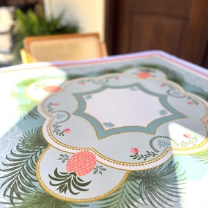 May include: A decorative table mat with a light blue background and a repeating pattern. The mat features a central design with a light blue border, a white interior, and a gold trim. Pink pineapples and green palm fronds are also featured.