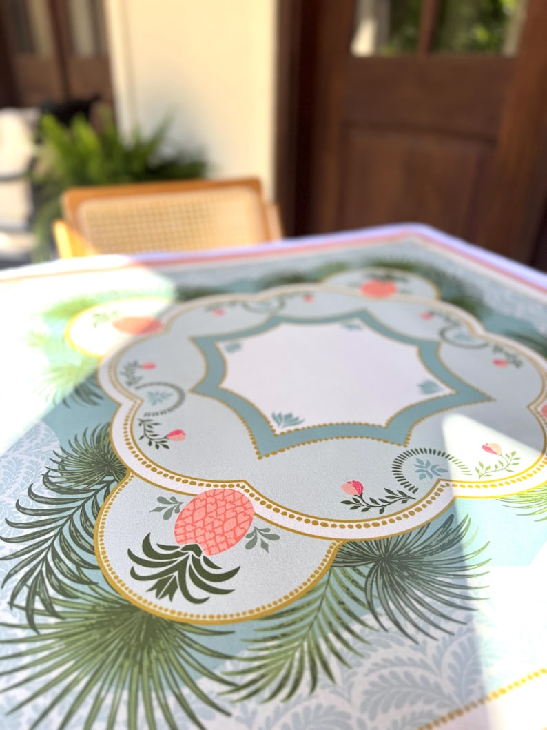 May include: A decorative table mat with a light blue background and a repeating pattern. The mat features a central design with a light blue border, a white interior, and a gold trim. Pink pineapples and green palm fronds are also featured.