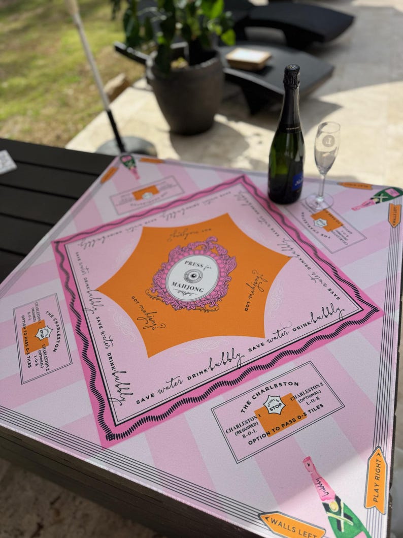 Pu&ograve; includere: Un tabellone di gioco del Mahjong rosa e arancione con la scritta "Press Mahjong" e "Save water drink bubbly". Una bottiglia di champagne verde e un calice da champagne sono sul tabellone. Il tabellone ha un motivo a strisce e le istruzioni del gioco.