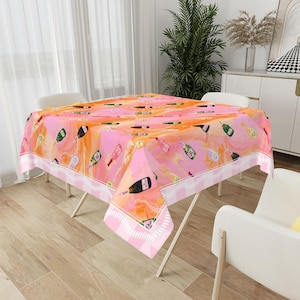 May include: A pink and orange tablecloth with a champagne bottle pattern. The tablecloth has a white and pink striped border and is set on a folding table. The background includes a window, chairs, and decorative items.