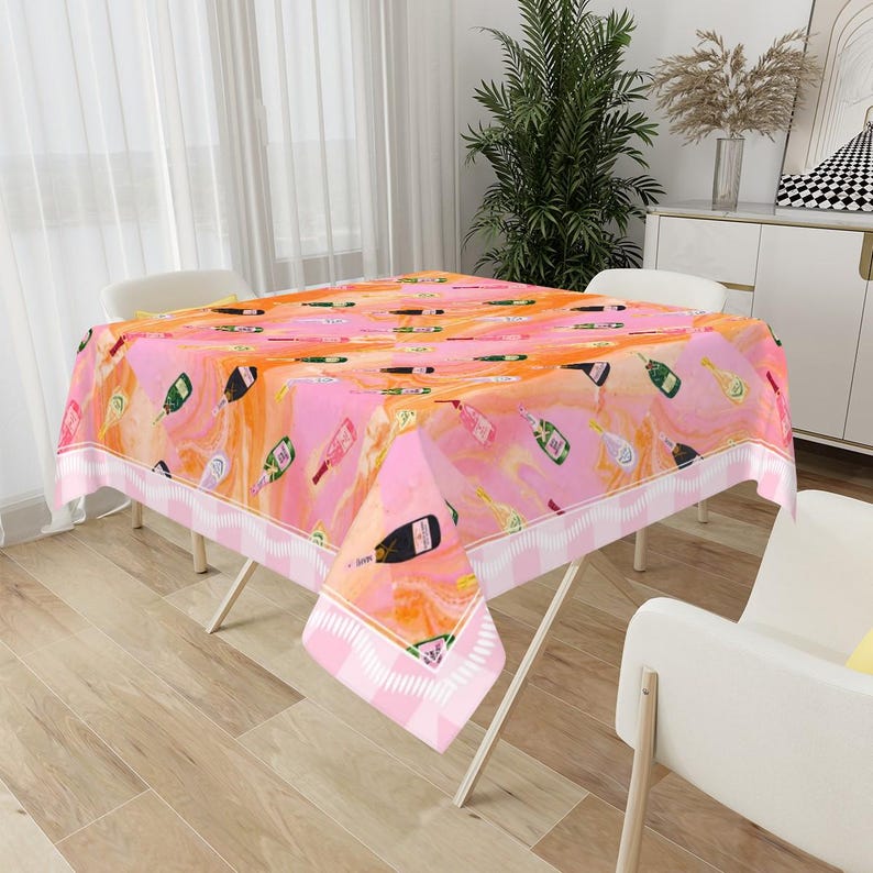 May include: A pink and orange tablecloth with a champagne bottle pattern. The tablecloth has a white and pink striped border and is set on a folding table. The background includes a window, chairs, and decorative items.