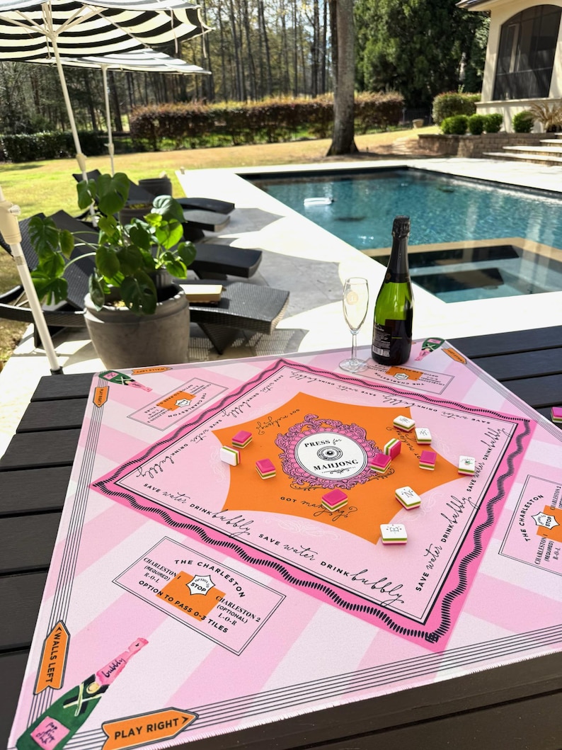 Pu&ograve; includere: Un tabellone di gioco del Mahjong rosa e arancione con il testo "Press for Mahjong" e "Save water drink bubbly." Una bottiglia di champagne e un bicchiere di champagne sono sul tavolo. Il tabellone di gioco &egrave; su un tavolo nero vicino a una piscina.