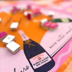 May include: A colorful illustration featuring a champagne bottle with the text "Bubbles & Bams Rose Imperial" and the word "Cheers". The design includes pink, orange, and black elements, with small, square objects scattered across the surface.