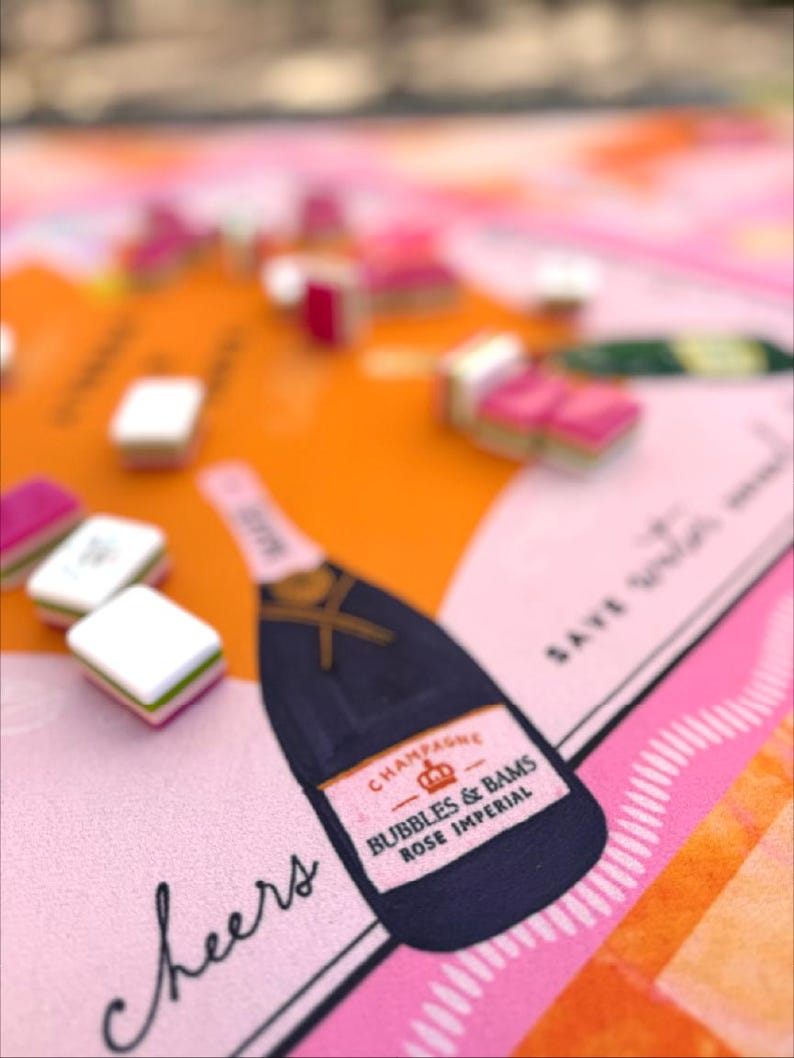 May include: A colorful illustration featuring a champagne bottle with the text "Bubbles & Bams Rose Imperial" and the word "Cheers". The design includes pink, orange, and black elements, with small, square objects scattered across the surface.