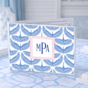 Puede incluir: Un portamenús de acrílico transparente con un estampado de grullas azules y blancas. El centro presenta un cuadrado rosa y blanco con las iniciales MPA en azul marino. El soporte está sobre un mantel azul claro estampado.