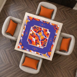 May include: A square game mat featuring a vibrant abstract pattern of orange, purple, and white shapes. The center is framed by a dark purple border with white text that reads THE CHANCELLOR and includes small icons. The mat sits on a light wood table.