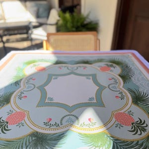 May include: A light blue tablecloth with a decorative border featuring pink pineapples, green palm fronds, and floral accents. The tablecloth has a central white space and a gold trim. The design is set against a light blue background.