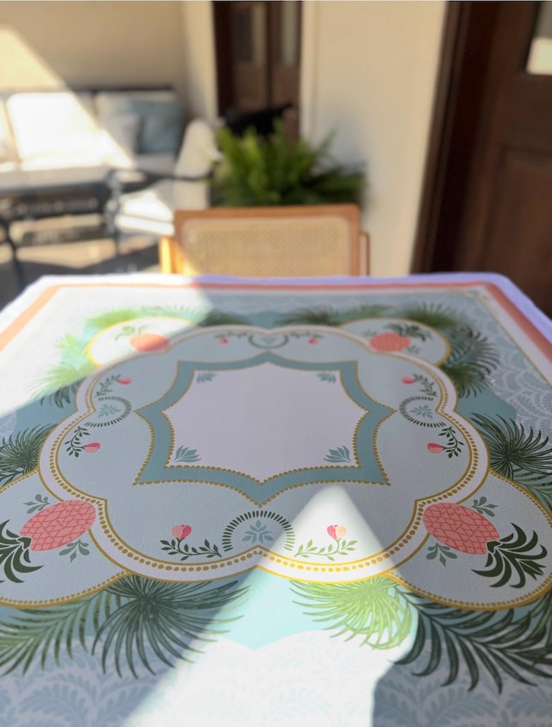 May include: A light blue tablecloth with a decorative border featuring pink pineapples, green palm fronds, and floral accents. The tablecloth has a central white space and a gold trim. The design is set against a light blue background.
