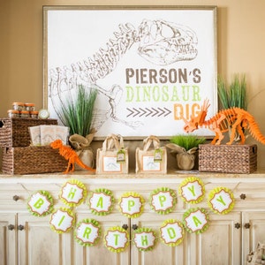May include: A dinosaur-themed birthday party setup with a sign that says "Pierson's Dinosaur Dig", a green plant, a dinosaur skeleton, and a banner that says "Happy Birthday".