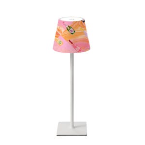 May include: A white table lamp with a square base and a tall, slender stem. The lampshade is a cone shape with a pink and orange abstract pattern featuring illustrations of champagne bottles. The lamp is designed for indoor use.