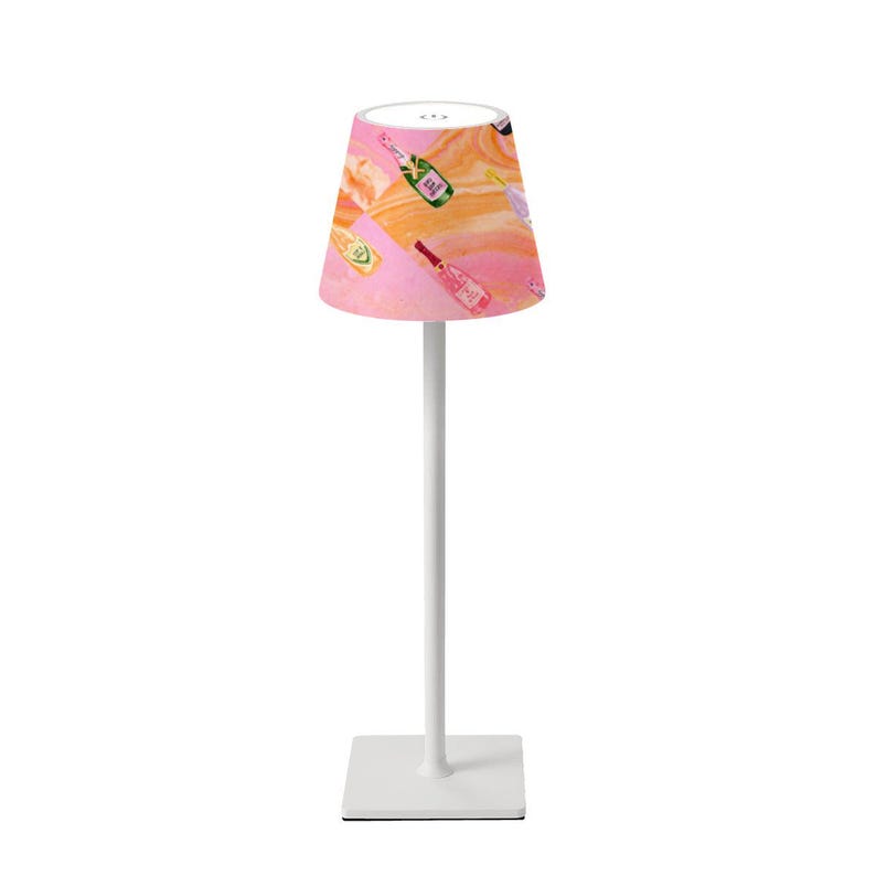 May include: A white table lamp with a square base and a tall, slender stem. The lampshade is a cone shape with a pink and orange abstract pattern featuring illustrations of champagne bottles. The lamp is designed for indoor use.