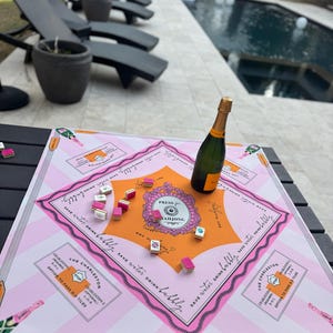 May include: A pink and orange Mahjong game board with a bottle of champagne. The board has the text "PRESS MAHJONG" and "SAVE WATER DRINK BUBBLY". Several Mahjong tiles are scattered on the board. The game is set up outdoors.