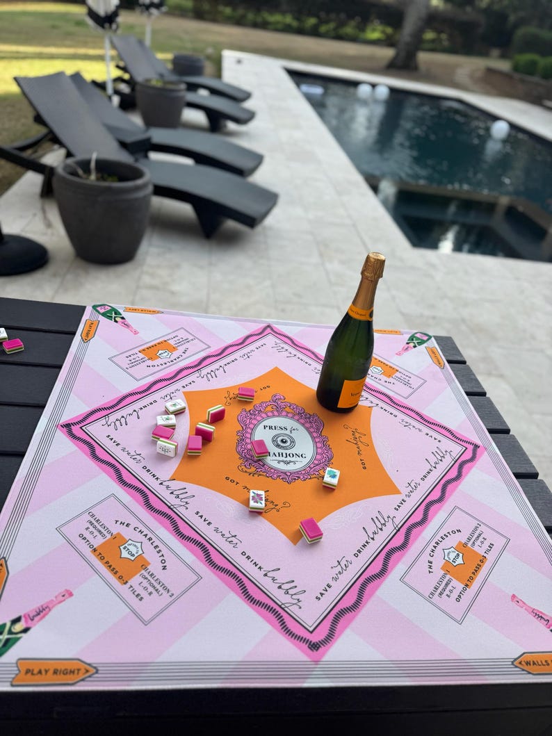May include: A pink and orange Mahjong game board with a bottle of champagne. The board has the text "PRESS MAHJONG" and "SAVE WATER DRINK BUBBLY". Several Mahjong tiles are scattered on the board. The game is set up outdoors.