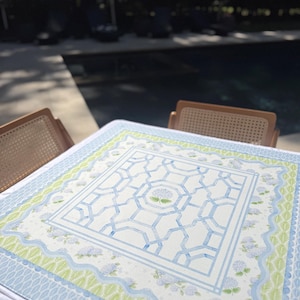 May include: A square tablecloth with a watercolor design. The central pattern features a blue geometric design with a floral motif. The border includes light blue, green, and white floral and geometric patterns. The tablecloth is set on a table with two wicker chairs.